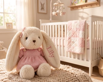 May include: A cream-colored plush rabbit toy with long ears, wearing a pink and white gingham dress with buttons. The name Lily is embroidered in pink thread on one ear. A small pink bow is attached to the top of the rabbit's head.