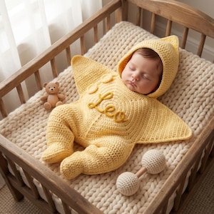 May include: A pale yellow crocheted star-shaped baby bunting with a hood and long sleeves. The garment features the name "Leo" embroidered in a cursive script on the chest. A small wooden teddy bear and a white crocheted rattle are nearby.