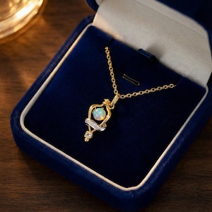 May include: A gold-toned necklace with a pendant featuring a round opal gemstone, accented with a diamond-studded silver band and a square-cut diamond. The necklace is presented in a blue velvet jewelry box, creating a luxurious feel.
