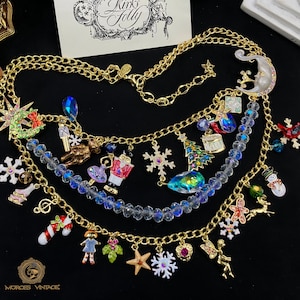 May include: A multi-strand gold-tone necklace with Christmas-themed charms, including a snowman, snowflakes, and a Christmas tree. The necklace also features a crescent moon pendant and a variety of colorful crystal beads. The brand name "Kirks Folly" is visible.
