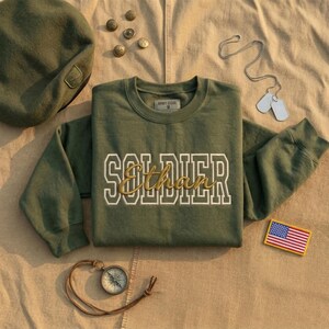 Puede incluir: Una sudadera verde oliva con la palabra "SOLDIER" en letras blancas y el nombre "Ethan" en escritura dorada. La imagen incluye una boina a juego, placas de identificación, una brújula y un parche de la bandera estadounidense.