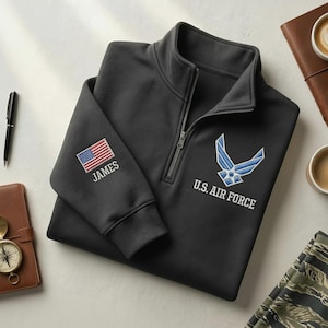 Könnte beinhalten: Ein anthrazitfarbenes Sweatshirt mit Viertelreißverschluss und dem Emblem und Text der US Air Force. Die amerikanische Flagge und der Name "JAMES" sind auf dem linken Ärmel gestickt. Ein Stift, ein Leder-Notizbuch und eine Kaffeetasse sind sichtbar.