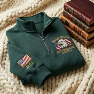Könnte beinhalten: Ein dunkelgrünes Sweatshirt mit Viertelreißverschluss und gestickten Details. Das Sweatshirt zeigt ein amerikanisches Flaggen- und Adlerdesign mit dem Text "UNITED STATES 250TH ANNIVERSARY 1776-2026". Der Ärmel hat eine gestickte amerikanische Flagge und den Namen "JACK".