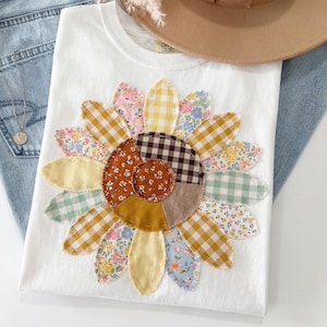 Może przedstawiać: Biała koszulka z dużym, aplikowanym wzorem słonecznika. Płatki słonecznika wykonane są z różnych wzorzystych tkanin, w tym kratki, nadruków kwiatowych i jednolitych kolorów. Koszulka ma okrągły dekolt i krótkie rękawy. Widoczny jest napis "Canvas Colors".