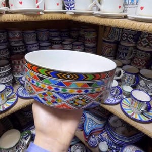 Puede incluir: Un cuenco grande de cerámica blanca con un colorido patrón geométrico en rojo, amarillo, azul y verde. El cuenco se sostiene frente a una estantería llena de diversos artículos de cerámica, incluyendo tazas, platillos y cuencos pequeños.