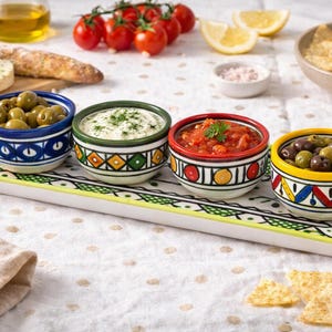 May include: A selection of appetisers presented on a serving tray. The tray holds four small, patterned ceramic bowls containing olives, dips, and salsa. Other items include tomatoes, lemons, and tortilla chips.