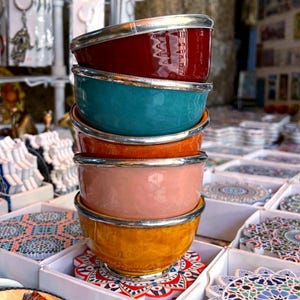 Puede incluir: Una pila de cuencos de cerámica coloridos con bordes plateados. Los cuencos son en tonos de rojo, turquesa, naranja, rosa y amarillo. Se exhiben sobre posavasos decorativos con patrones intrincados, mostrando una variedad de artículos de cocina hechos a mano.