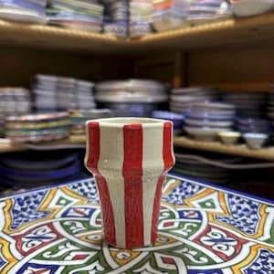 Puede incluir: Un vaso de cerámica con rayas verticales rojas y blancas. El vaso está sobre un plato cuadrado decorativo con un patrón geométrico colorido. Estantes en el fondo muestran pilas de cuencos y platos de cerámica.