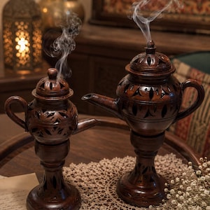 May include: Two dark brown, carved wooden incense burners with smoke rising from the tops. The burners have intricate cut-out designs and are placed on a wooden tray with a lace doily and dried flowers.