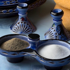 Può includere: Set di sale e pepe in ceramica blu con portacandele abbinati. Il piatto per sale e pepe è diviso in due sezioni, una riempita di pepe nero e l'altra di sale bianco. I portacandele sono blu con accenti neri e motivi decorativi.