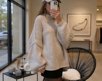 May include: A beige, oversized turtleneck sweater with long, voluminous sleeves. The sweater is paired with a gray baseball cap and a black skirt. The image is taken indoors, with a modern aesthetic.