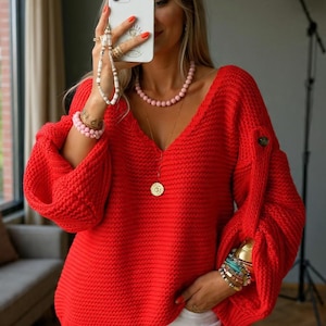 May include: A bright red, chunky knit sweater with a deep V-neck and long, voluminous sleeves. The sweater is paired with white pants. The person is wearing a pink beaded necklace and bracelets, and holding a white phone with a strap.