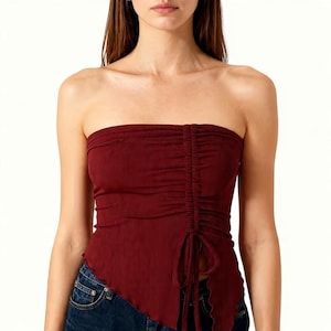 May include: A burgundy tube top with a ruched front and a drawstring detail. The top has a ribbed texture and an asymmetrical hemline. The model is wearing blue jeans.