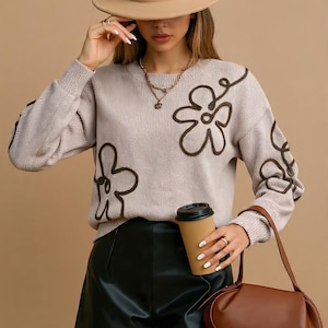 May include: A beige sweater with brown floral designs, paired with black shorts, a brown purse, and a beige hat. The outfit includes a coffee cup and gold necklaces. The sweater features a crew neck and long sleeves.
