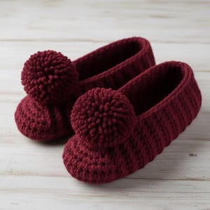 以下が含まれることがあります： 手作りの、バーガンディ色の編み物スリッパのペア。各スリッパは、上部に大きなポンポンとリブ編みのデザインが特徴です。スリッパは明るい色の木製の表面に展示されています。
