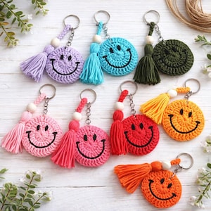 May include: A collection of handmade crochet smiley face keychains in various colors, including purple, blue, green, pink, red, and orange. Each keychain features a tassel and a metal ring for attaching keys.