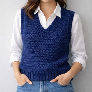 Può includere: Un gilet blu navy lavorato a mano con scollo a V. Il gilet è indossato sopra una camicia bianca con colletto e jeans blu. La trama a maglia è visibile e il gilet sembra essere fatto di un filato morbido. La persona indossa il gilet con le mani in tasca.