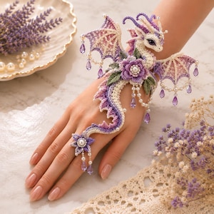 May include: A crochet dragon hand ornament featuring a gradient of purple, pink, and cream yarn. The design includes intricate wings, a central purple flower with a pearl center, dangling purple crystal teardrops, and small white pearls.