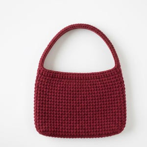 May include: A burgundy crochet handbag with a rounded shape and a single handle. The bag is made of a textured, knitted material and has a simple, classic design. The handle is arched and attached to the top of the bag.