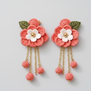 May include: Pair of coral-colored crochet flower earrings. Each earring features a flower with white and gold accents, green leaf, and dangling chains with coral-colored ball charms. The earrings are handmade.