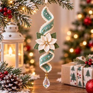Puede incluir: Un adorno navideño colgante con una cinta en espiral de color verde salvia decorada con pequeñas perlas blancas y cuentas doradas. Una flor de pascua blanca con hojas verdes se sitúa en el centro, con un cristal transparente en forma de gota.