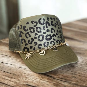 Puede incluir: Gorra de camionero verde oliva con un diseño de estampado de leopardo en el panel frontal. La gorra presenta una cadena dorada con dijes, incluyendo un lazo, un corazón y una carita sonriente. La parte trasera de malla y la visera son de color verde oliva.