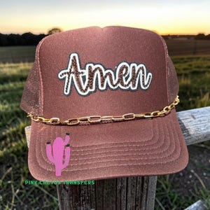 May include: Brown trucker hat with the word "Amen" in glittery letters. A gold chain is attached to the brim. A pink cactus graphic is on the front. The hat is sitting on a wooden post.