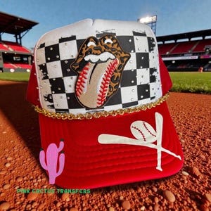 Puede incluir: Gorra de béisbol roja y blanca con un patrón de tablero de ajedrez y un logotipo de lengua con temática de béisbol. La gorra presenta un diseño de béisbol y bates, un ribete de cadena dorada y un cactus rosa. El texto "PINK CACTUS TRANSFERS" es visible.