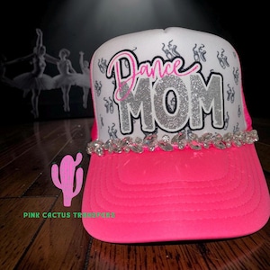 May include: A white and cerise baseball cap with the words "Dance MOM" in pink and silver glitter. The cap has a cerise brim and is decorated with rhinestones and ballet shoes.