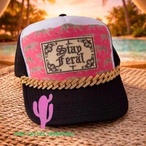 May include: Black trucker hat with a white top panel and pink accents. The hat features a gold chain across the brim and a pink cactus graphic. The front panel displays the text "Stay Feral" in a decorative frame.