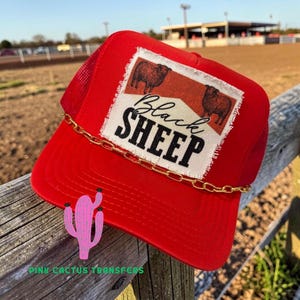May include: A bright red trucker hat with a mesh back and a gold chain detail. The front patch displays the words "Black Sheep" and sheep illustrations. A striking accessory.