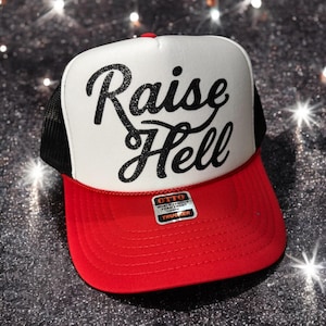 May include: A red, white, and black trucker hat with the words "Raise Hell" in glittery black script across the front. The hat has a red brim, a white front panel, and a black mesh back. A small tag is attached to the brim.