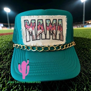 Puede incluir: Gorra de camionero color turquesa con detalle de cadena dorada y un parche que dice "MAMA" en una fuente colorida y goteante. Un gráfico de cactus rosa está en la parte delantera. El texto "PINK CACTUS TRANSFERS" está en la parte inferior.