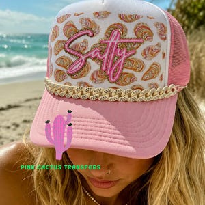 May include: A pink and white trucker hat featuring a repeating oyster shell pattern on the front panel. The word Salty is written in pink script with a gold glitter outline. A cream-colored chain link trim sits above the pink brim.
