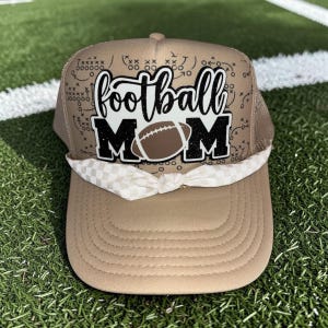 May include: Tan trucker hat with the words "football MOM" in black and white, featuring a football graphic. The hat has a white and tan checkered bow and a mesh back.