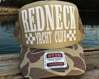 May include: A trucker hat with a tan front panel and a camouflage pattern on the bill. The hat features the text "REDNECK YACHT CLUB" in white block letters. The hat has a mesh back and a snap closure.
