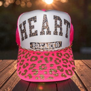 May include: A pink trucker hat with a leopard print design and the words "HEART BREAKER" in large, bold letters. The hat features a white front panel, a pink mesh back, and a gold chain detail.
