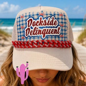 Puede incluir: Una gorra tipo camionero beige con un patrón de cuadros vichy azul y blanco en el panel frontal y pequeños cangrejos rojos. El texto Dockside Delinquent está escrito en purpurina roja. Una cadena roja con pedrería adorna la base.