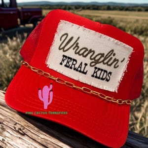 May include: Red trucker hat with a white patch that reads "Wranglin' FERAL KIDS" in black script. A gold chain accents the brim, and a pink cactus graphic is on the front. The text "PINK CACTUS TRANSFERS" is printed on the brim.