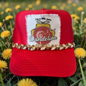 May include: A red trucker hat with a mesh back and a decorative jeweled band. The front patch features a dandelion design with the text "I'm a little more Dandelion."
