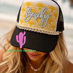 May include: A black and white trucker hat featuring a yellow sunburst pattern on the front panel. The word Beachy is written in silver glitter script across the front. A thick, silver-toned chain trim sits above the black brim, which displays a pink cactus graphic and the text PINK CACTUS TRANSFERS.