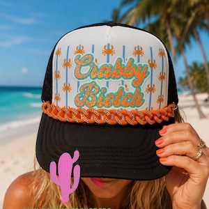 Puede incluir: Una gorra tipo camionero en blanco y negro con un panel frontal blanco que presenta un patrón repetitivo de pequeños cangrejos naranjas y el texto Crabby Biotch en purpurina naranja. Incluye una cadena naranja y una visera negra.