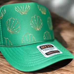 May include: Green trucker hat with gold glitter seashell pattern. The hat features a curved brim, a mesh back, and a snap closure. The hat has a tag that says "OTTO WORLD FAMOUS FOAM FRONT TRUCKER".