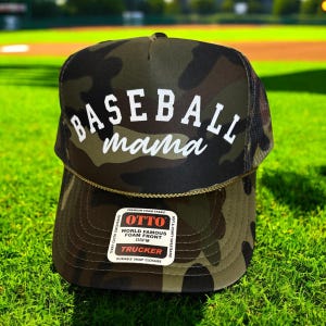 May include: A camouflage trucker hat with the words "BASEBALL mama" in white script. The hat has a mesh back and a snap closure. The hat is sitting on green grass.