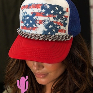 May include: A red, white, and blue trucker hat with an American flag design on the front panel. The hat features a red brim, a white front panel with a distressed flag print, a blue mesh back, and a silver chain detail.
