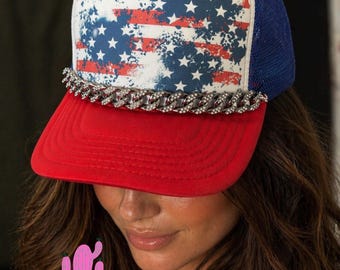 May include: A red, white, and blue trucker hat with an American flag design on the front panel. The hat features a red brim, a white front panel with a distressed flag print, a blue mesh back, and a silver chain detail.