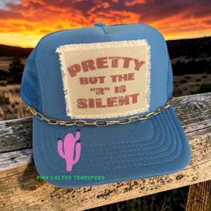 May include: Blue trucker hat with a mesh back and a curved brim. The hat features a patch with the text "PRETTY BUT THE "R" IS SILENT" in brown letters. A pink cactus graphic is on the front, and a gold chain is attached to the brim.
