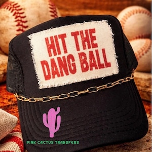 May include: Black trucker hat with a gold chain and a pink cactus graphic. The hat features the text "HIT THE DANG BALL" in red letters on a white patch. The words "PINK CACTUS TRANSFERS" are printed in green at the bottom of the hat.