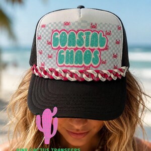 May include: A black and white trucker hat featuring a checkered pattern with small red crab graphics. The front panel displays the text "COASTAL CHAOS" in glittery teal letters. A decorative chain link trim in alternating pink and white sits above the brim.