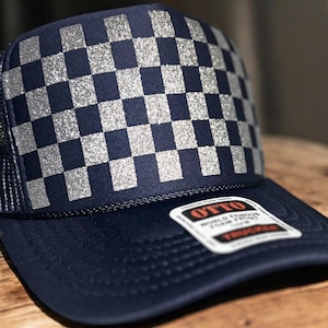 May include: Navy blue trucker hat with a silver glitter checkerboard pattern on the front panel. The hat has a mesh back and a curved brim. A white label with black text is attached to the brim.